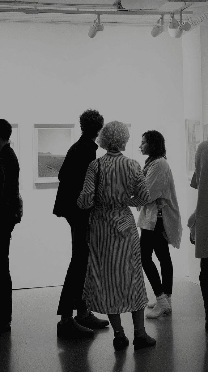 Black and white photograph of visitors viewing framed artwork in a gallery space with track lighting. A person in a striped dress stands prominently in the center, surrounded by other gallery visitors examining the wall-mounted pieces. (AI-generated image) by Sydney SG