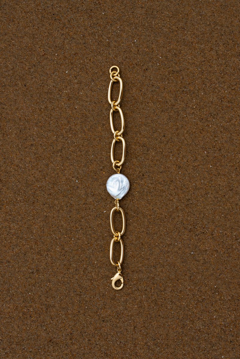 Single gold chain bracelet with a white pearl laid flat on brown sand. The simple, clean composition highlights the contrast between the metal and natural elements. by Sydney SG