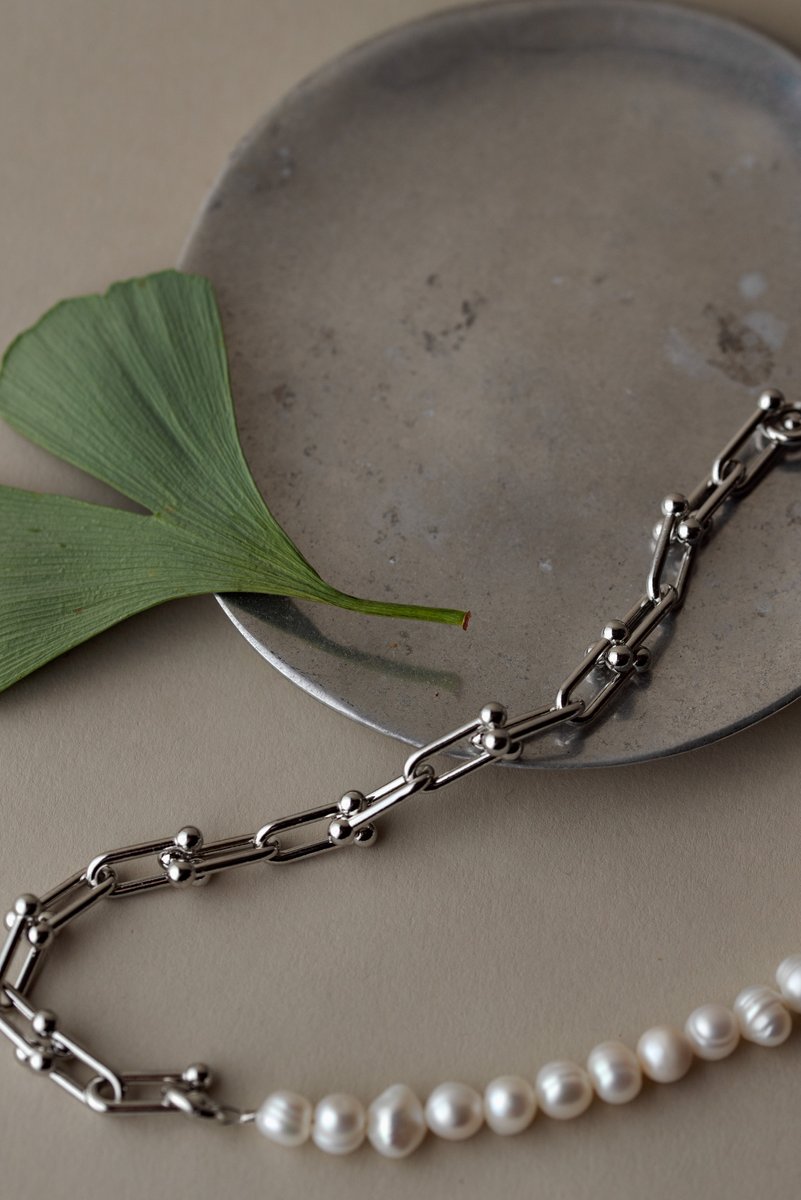 Silver chain bracelet with pearls arranged next to a green ginkgo leaf and a textured silver dish on a neutral background. An elegant composition of jewelry and nature. by Sydney SG