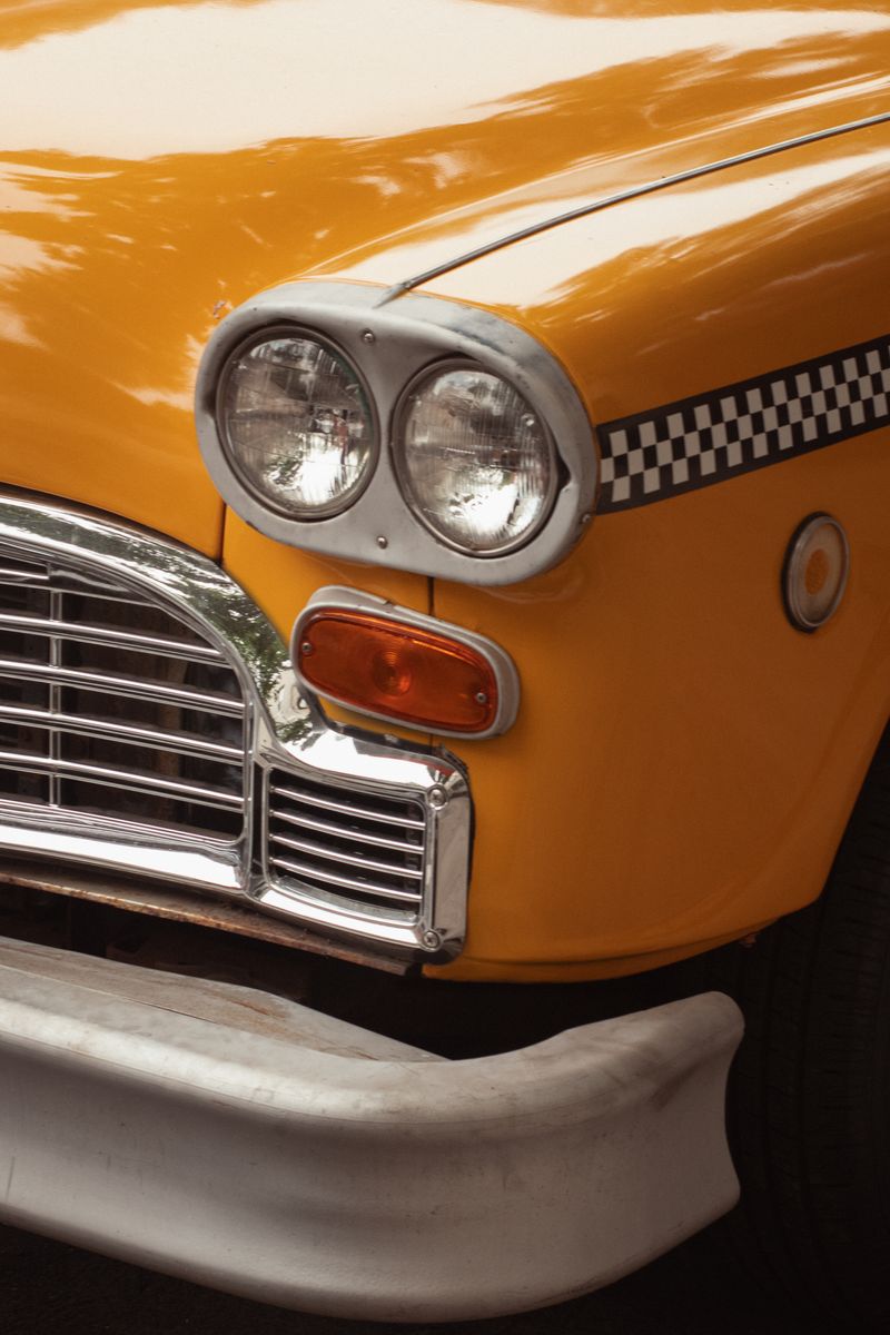 Close-up detail of a vintage yellow taxi cab showing dual headlights, chrome grille, and checkered stripe pattern. The bright orange-yellow paint and classic automotive details capture iconic urban transportation. by Sydney SG