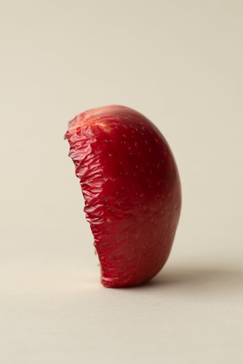 A halved red apple that has begun to rot, with wrinkled skin against a beige background. by Sydney SG