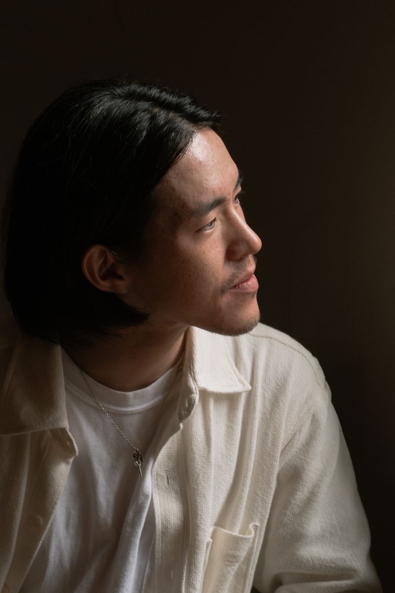 Profile portrait of a person in a white collared shirt against a dark background, lit with dramatic side lighting. The warm illumination creates strong contrast and highlights the subject's contemplative expression. by Sydney SG