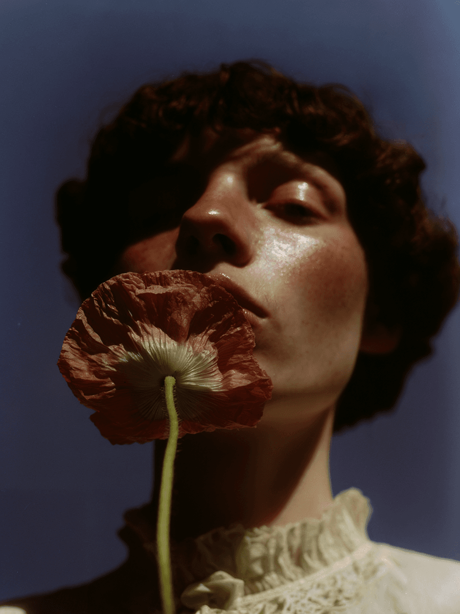 Portrait of a person with curly dark hair tilted back with eyes closed, holding a deep red poppy flower near their face. The dramatic blue lighting and the flower's rich burgundy color create a striking artistic composition. (AI-generated image) by Sydney SG