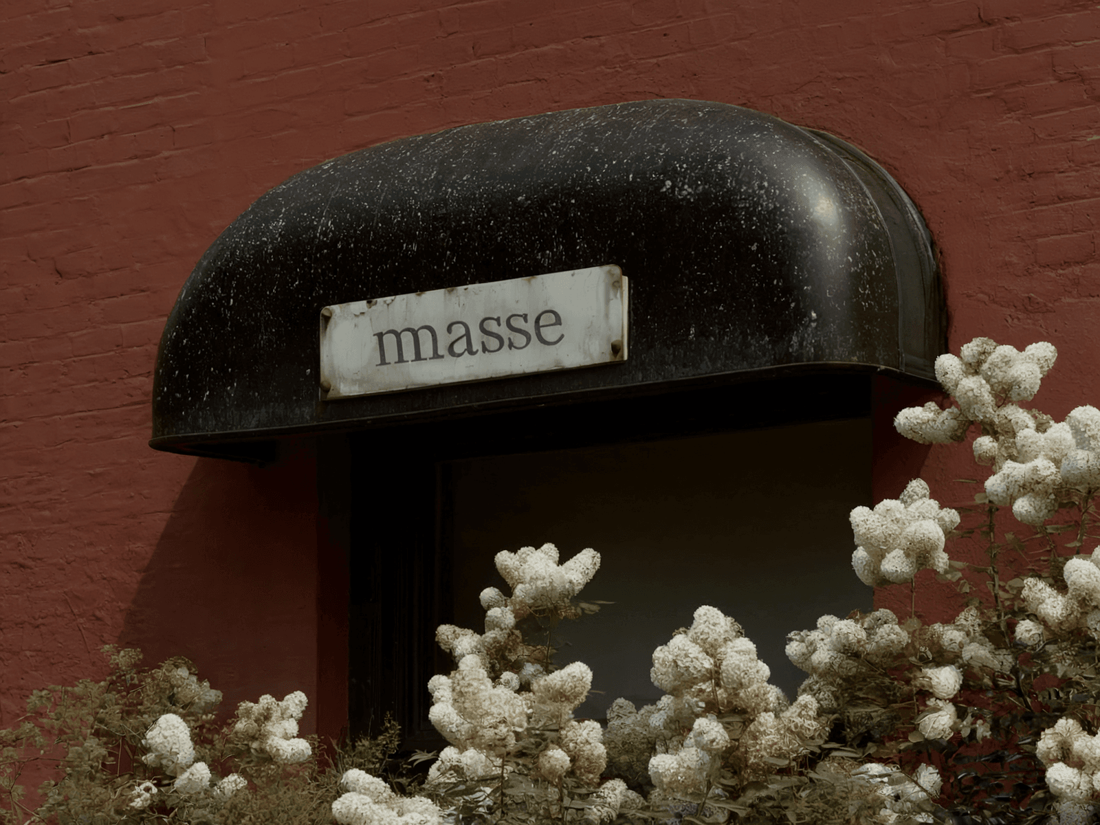 A black curved storefront awning with "masse" signage above white hydrangea flowers. The awning contrasts against red brick walls, creating a striking architectural detail with natural elements. (AI-generated image) by Sydney SG