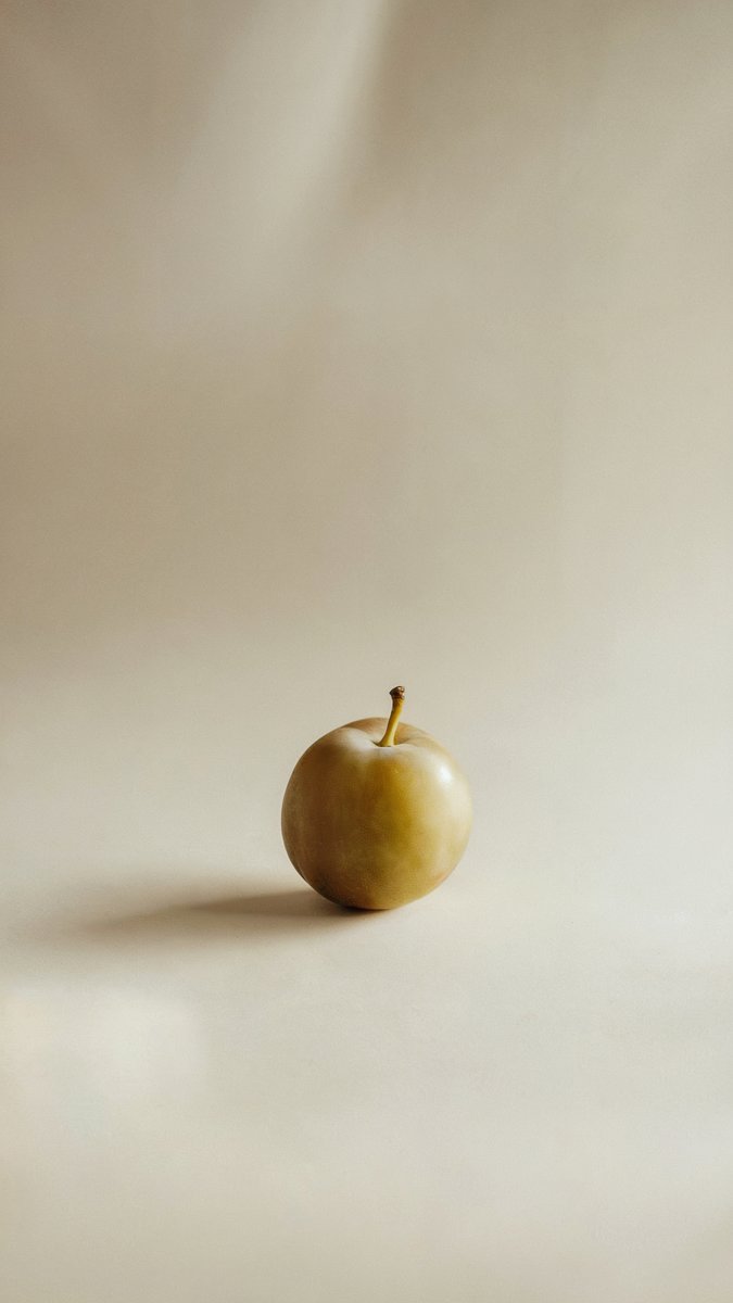 A single golden-green plum on a neutral beige background with soft lighting casting a faint shadow. Image generated with AI. by Sydney SG