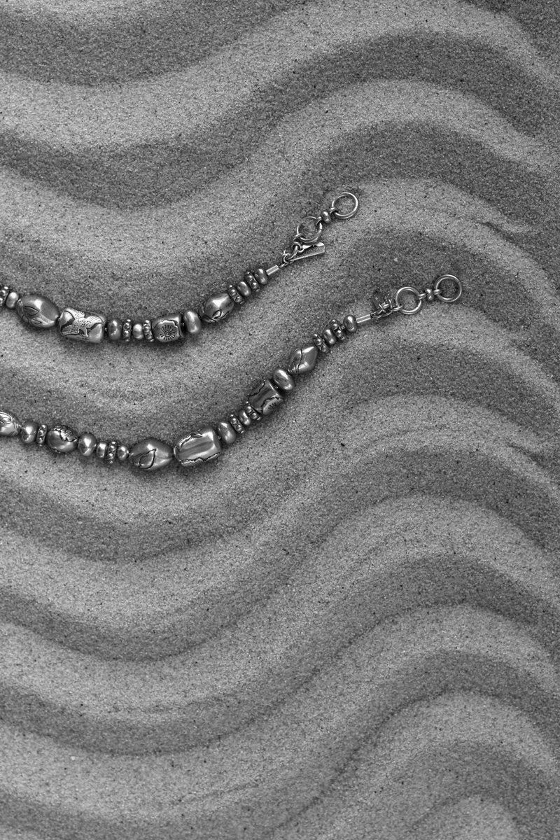 Two silver beaded bracelets lying on textured, wavy sand patterns, captured in black and white. by Sydney SG