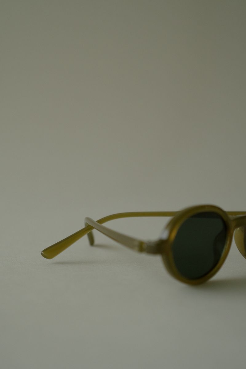 Close-up of chartreuse sunglasses resting on their side, revealing the sculptural temple and lens curvature. The warm translucent material absorbs and filters light, creating depth within the frame against the soft, diffused background. by Sydney SG