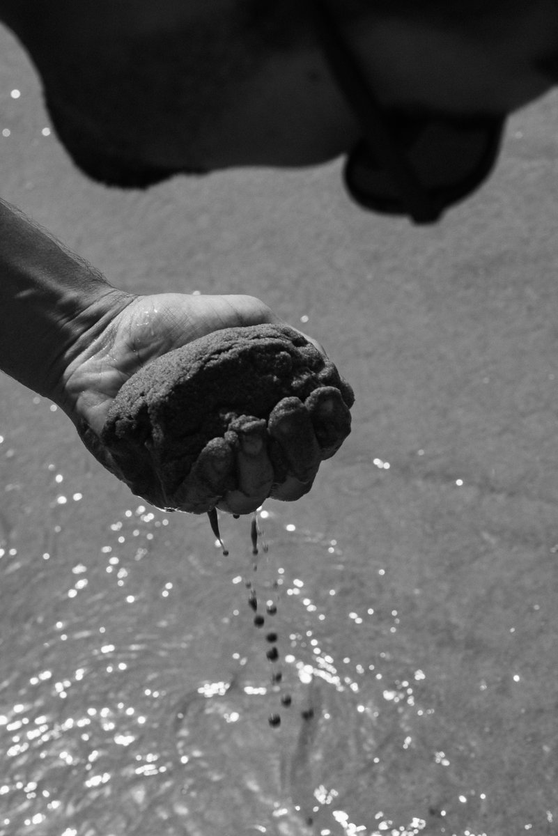 Close-up of a hand holding wet sand over a body of water. The sand is slowly dripping from the hand, capturing a moment of interaction with the natural elements. by Sydney SG