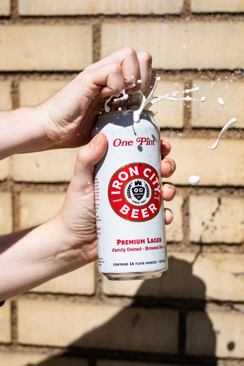 Hand tightly gripping a can of "Iron City Beer," with foam spraying outward against a brick wall background in sunlight. by Sydney SG