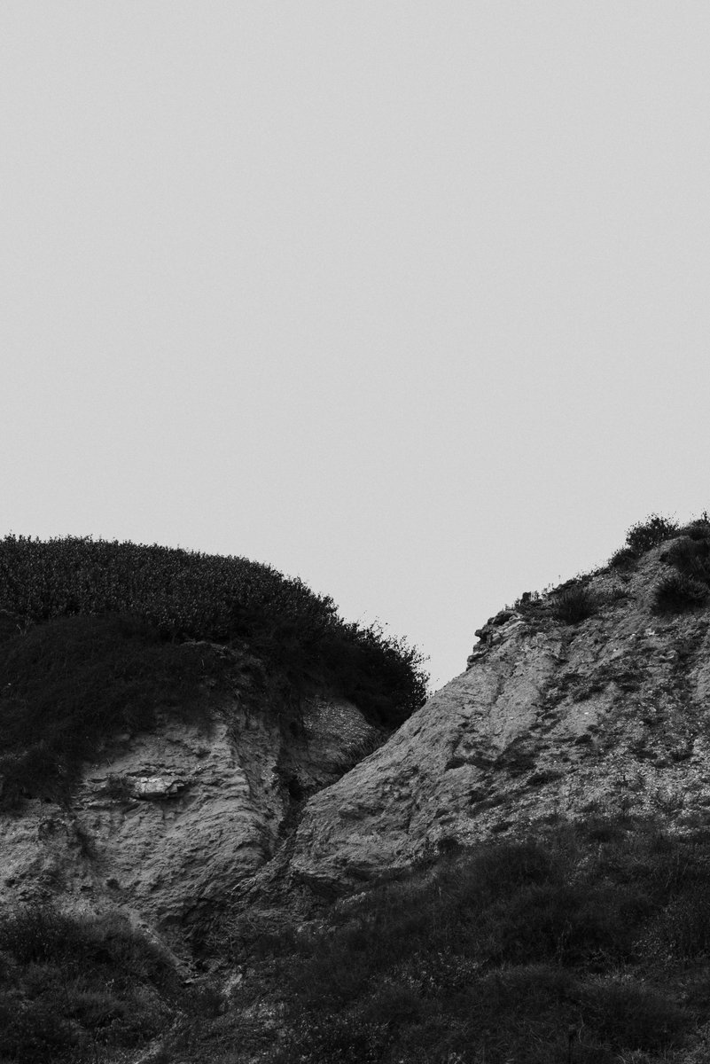 Black and white photograph of two rocky hills separated by a narrow gap, covered with sparse vegetation. A minimalist landscape evoking a moody and stark atmosphere. by Sydney SG