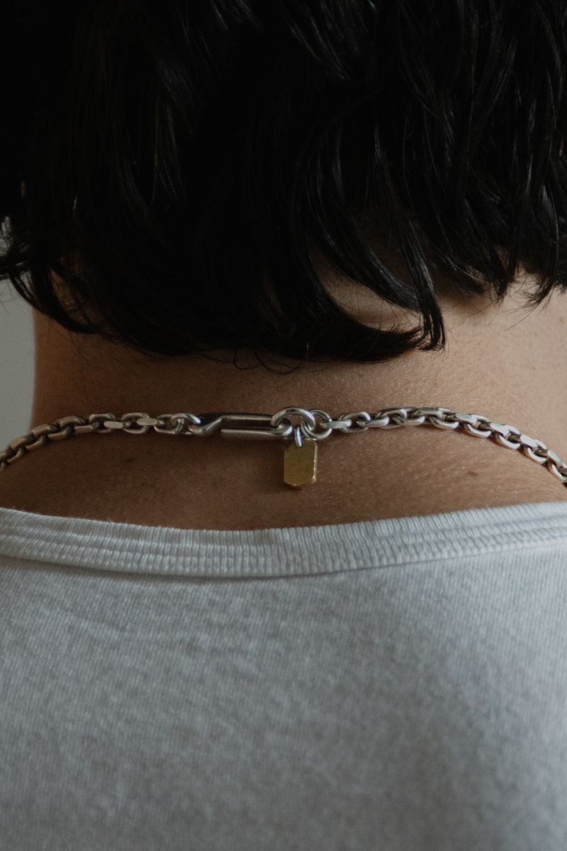 Closeup of a silver chain necklace with small pendant resting against the nape of the neck. The contrast between metal, skin tone, and white fabric creates a subtle study in texture and personal adornment. by Sydney SG