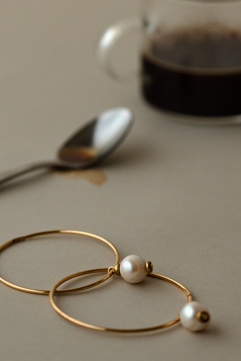 Two gold hoop earrings with pearls arranged on a neutral surface next to a glass cup of espresso and a spoon. A lifestyle shot with a quotidian feel. by Sydney SG