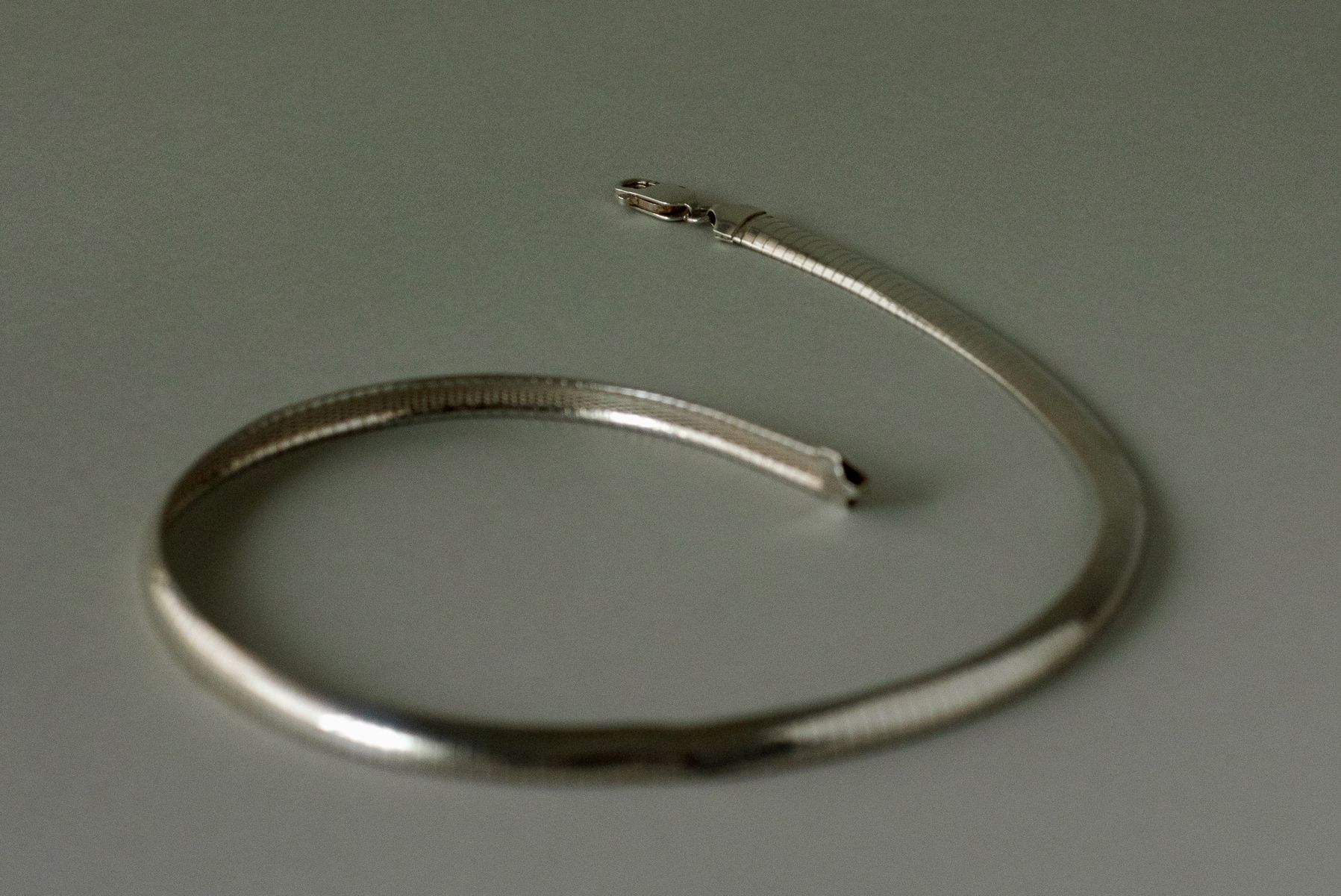 Minimalist silver snake chain bracelet coiled in a loose spiral on a muted gray-green surface. The fine ribbed texture catches light along its sleek, flexible body, emphasizing the piece's sinuous movement and the technical precision of its construction. by Sydney SG