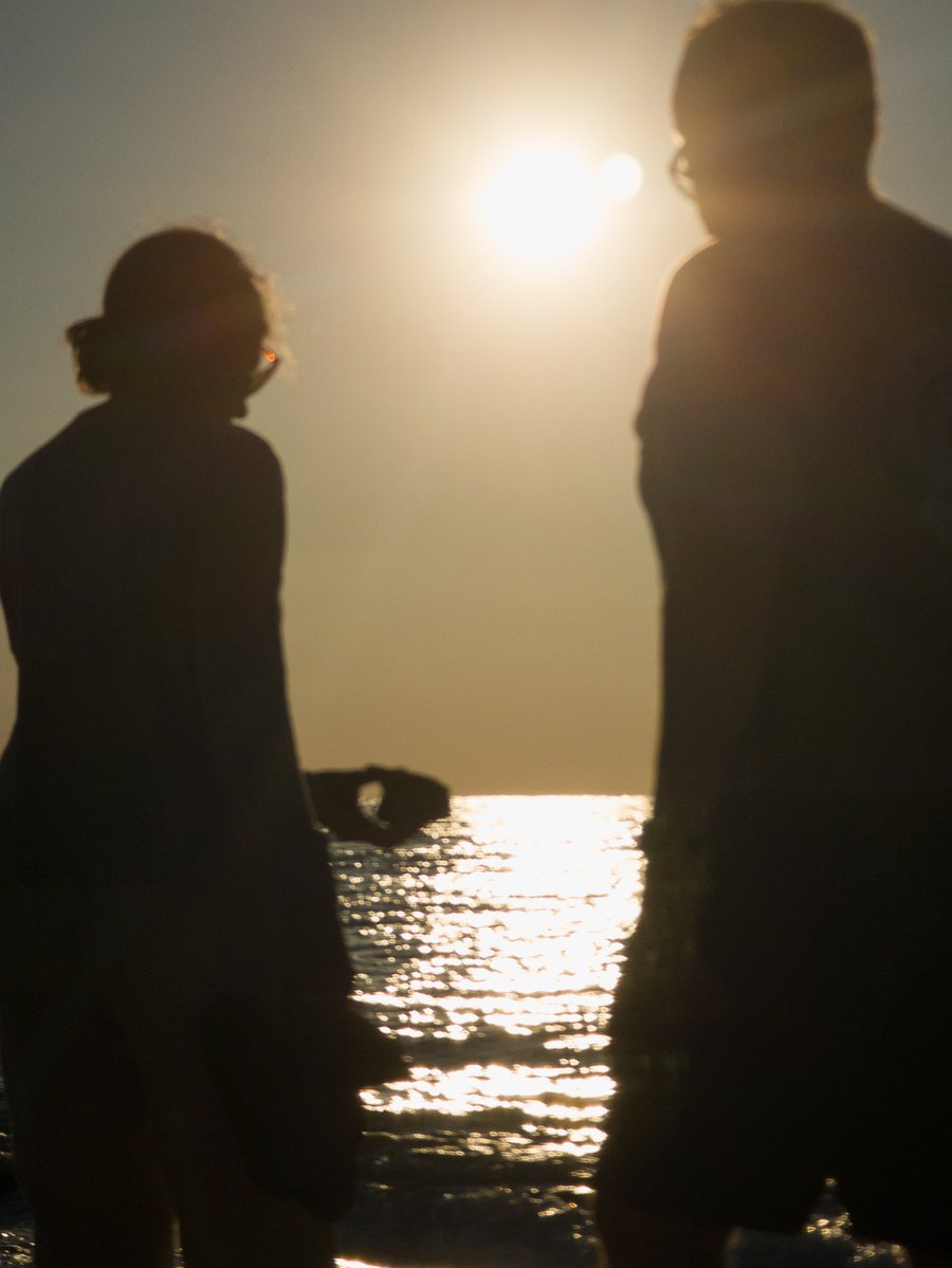 Two shadowed figures, one holding an item, walking by a sunlit ocean. The sun sets low in the sky, casting a warm glow over the scene. by Sydney SG