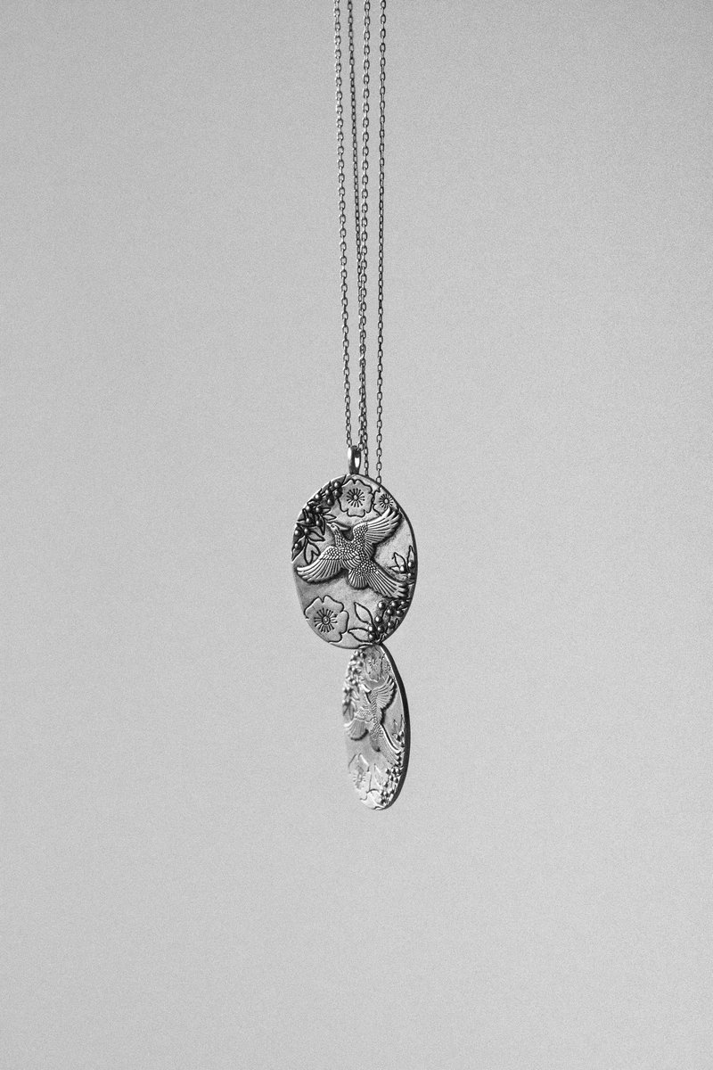 A silver pendant featuring floral and bird engravings, hanging from thin chains against a plain gray backdrop. by Sydney SG