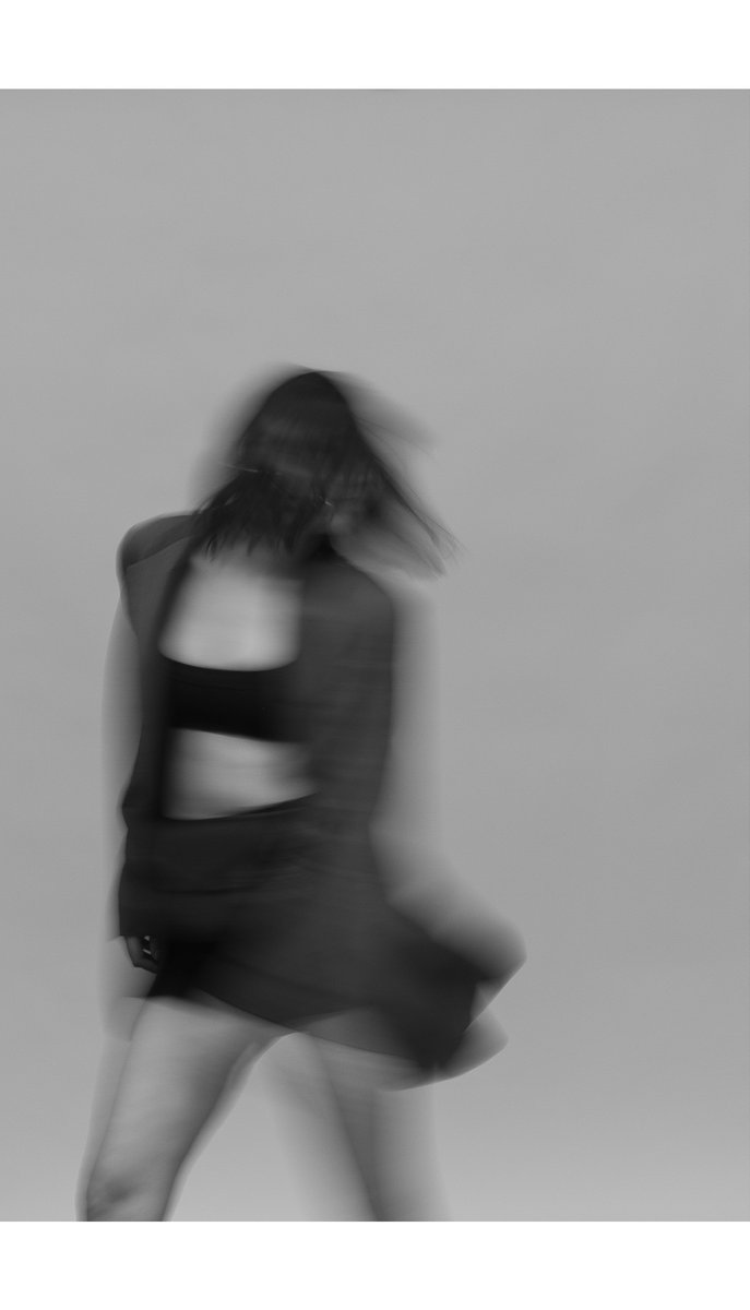 Black and white image of a woman in motion, wearing a cropped blazer and skirt, creating a sense of movement. by Sydney SG