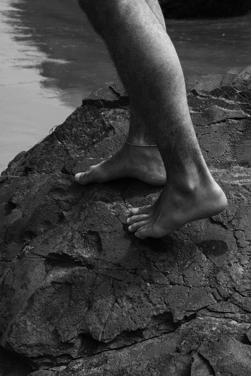 Black and white photo of bare feet walking across rough, rocky terrain near the water. The texture of the skin and rock surface is accentuated by the stark lighting. by Sydney SG