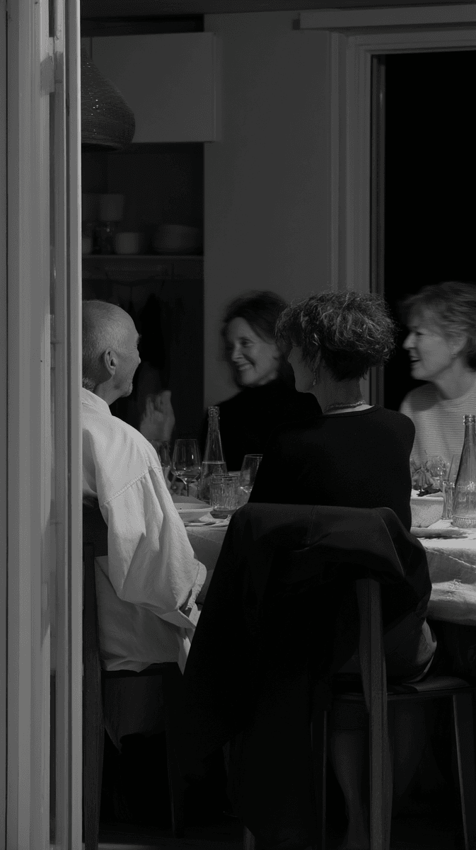 Black and white photograph of people gathered around a dining table in conversation, with wine glasses and bottles visible. The intimate scene captures a moment of social connection in what appears to be a home dining room. (AI-generated image) by Sydney SG