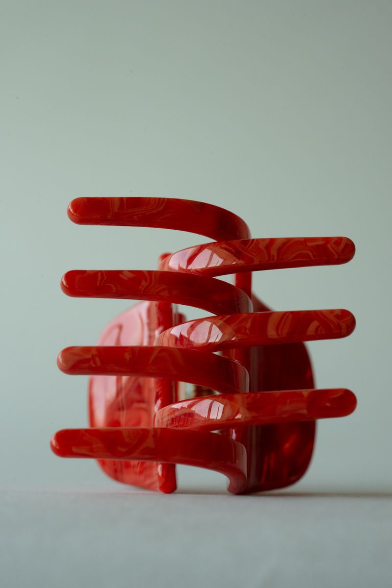 A sculptural red translucent hair claw standing vertically, creating rhythmic negative space between each glossy claw tooth. The marbled crimson material catches light along its edges, conveying both fluidity and architectural precision. by Sydney SG