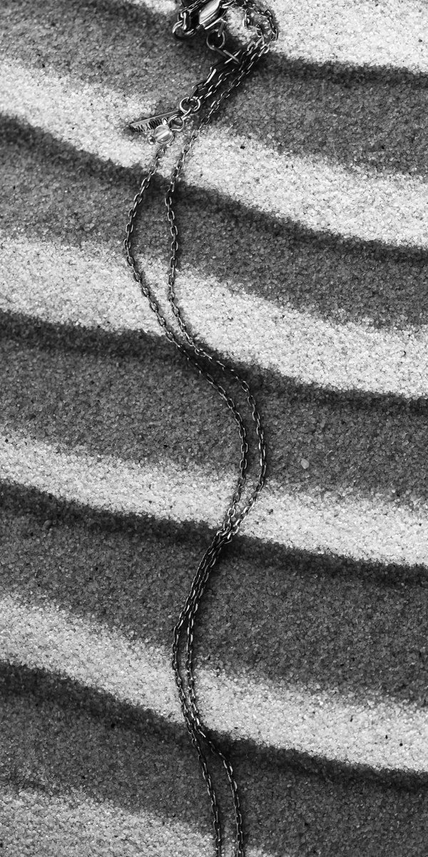 Black and white photo of a metal chain resting on wavy, textured sand patterns, with the chain trailing diagonally. by Sydney SG