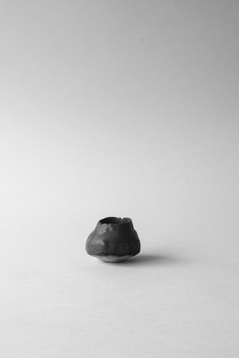 Small, irregularly shaped ceramic bud vase, rough in texture, placed on a neutral surface with lots of negative space. by Sydney SG