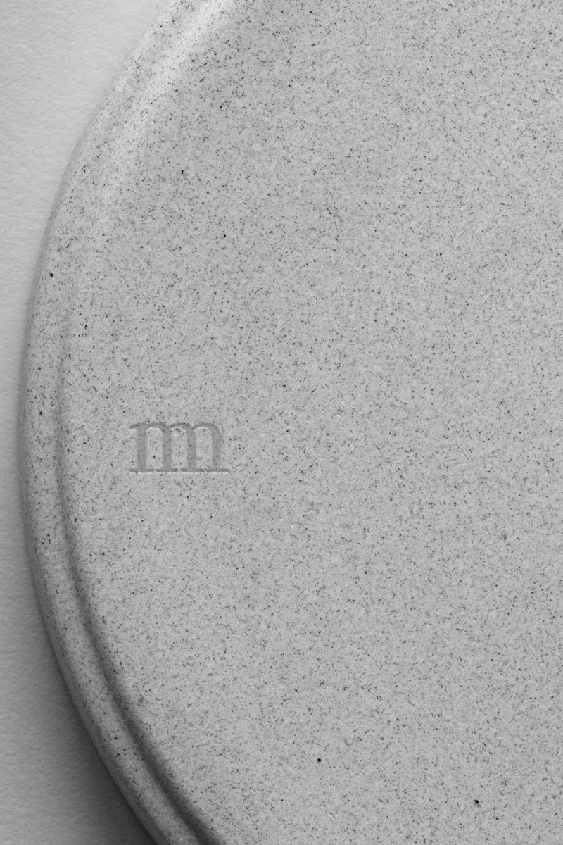 Close-up of a round, textured ceramic surface, neutral in tone, with the letters "m" subtly engraved. by Sydney SG