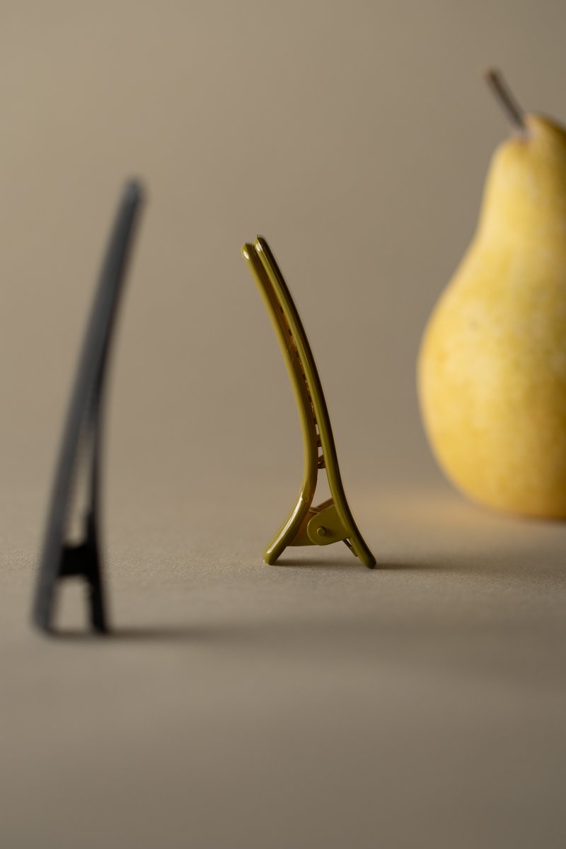A shallow depth of field still life composition with a black, long-nosed hair clip in the foreground, a similar green hair clip centered and in focus, and a bright yellow pear in the background, all standing upright on beige paper. by Sydney SG