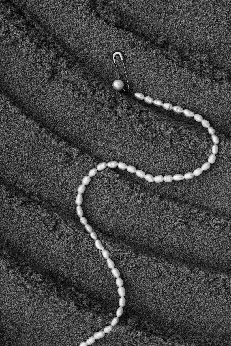 Pearl necklace arranged in a sinuous line across textured sand, with subtle shadows creating a sense of movement. A mix of natural and delicate elements in a monochromatic palette. by Sydney SG