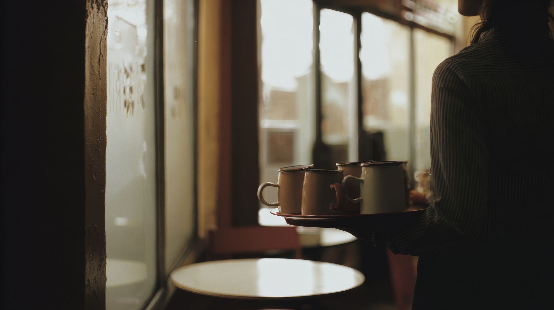 A server carries a tray of enamel mugs through a cafe with tall windows with natural light streaming in. The warm golden tones create an intimate café atmosphere with a person visible in dark clothing. (AI-generated image) by Sydney SG