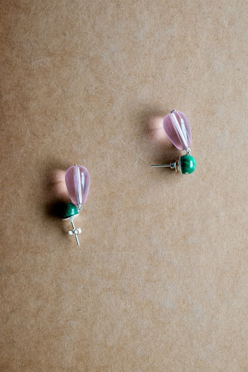 A pair of earrings with green stone studs and translucent pink teardrop dangles arranged on a beige textured surface. The jewelry displays elegant color contrast between the emerald green and soft lavender tones. by Sydney SG