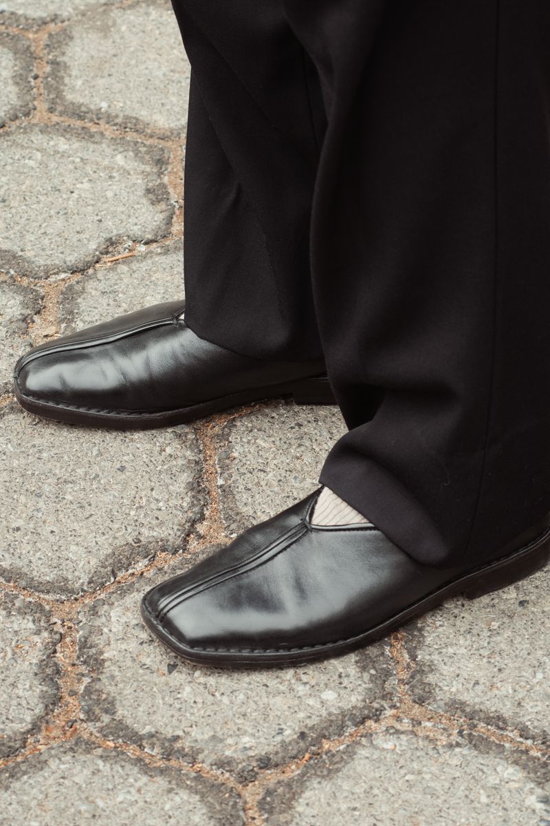 Black leather Lemaire shoes and dark pants photographed from above on cracked concrete pavement. The formal footwear contrasts with the weathered, textured urban surface beneath. by Sydney SG