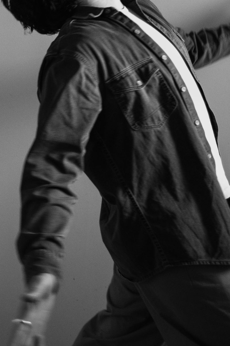 Motion-blurred figure in dark denim shirt and white undershirt, captured mid-dance. The monochromatic treatment and intentional blur transform an ordinary moment into a study of form and gesture. by Sydney SG