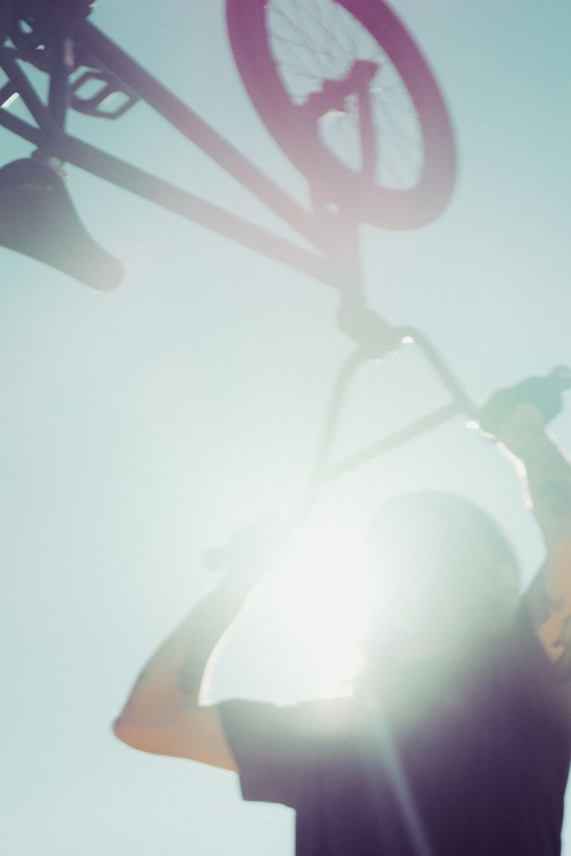 An abstract image of a BMX rider swinging his bike upwards towards the sky. The sun glares down through the handlebars creating a multicolor flare across the image. by Sydney SG