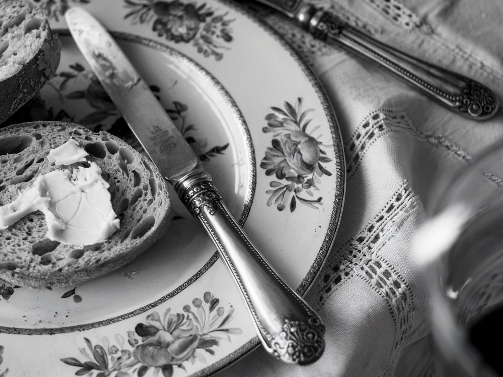 Black and white close-up of a slice of bread with butter on a vintage floral plate. A butter knife rests on the plate, creating a rustic, elegant dining scene. Image generated with AI. by Sydney SG