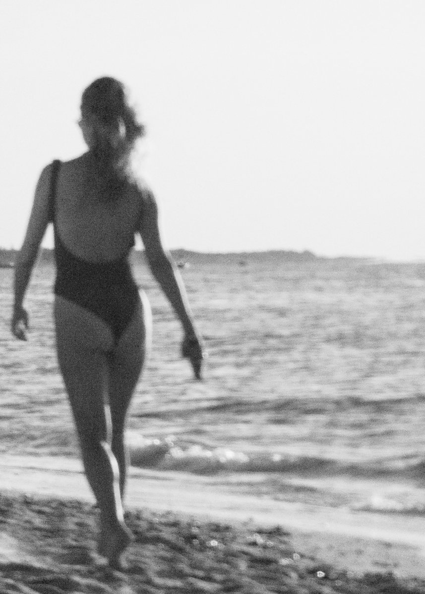 A grainy, black and white photo of a woman in a black, low-back, one-piece swimsuit walking away from the camera down the beach and towards the horizon. by Sydney SG