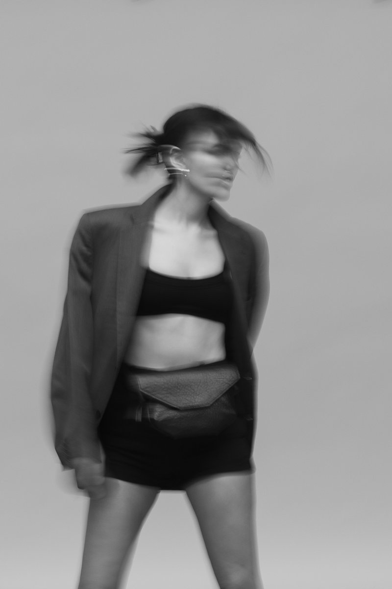 Blurred black and white portrait of a woman in motion, wearing a blazer, crop top, and a crossbody bag, with her hair moving dynamically. by Sydney SG