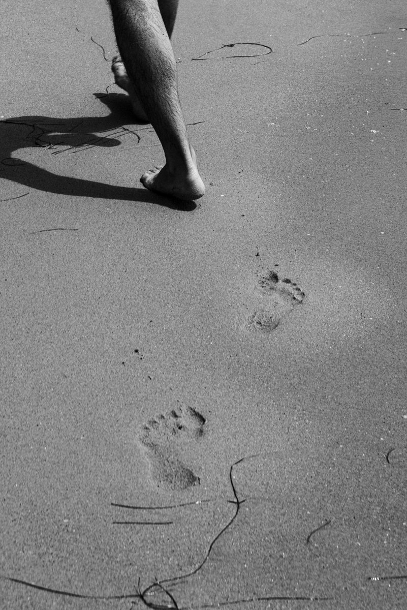 Black and white photo of a person walking barefoot on sand, leaving visible footprints behind. The shadow and texture of the sand add depth to the minimalist scene. by Sydney SG