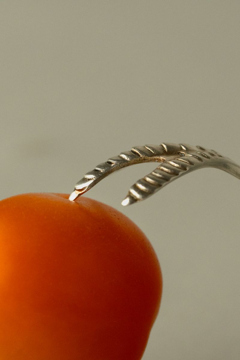Silver, claw-like fork tines piercing a vibrant orange-red cherry tomato against a minimal gray background. The composition highlights the contrast between the polished metal and the fruit's natural form. by Sydney SG