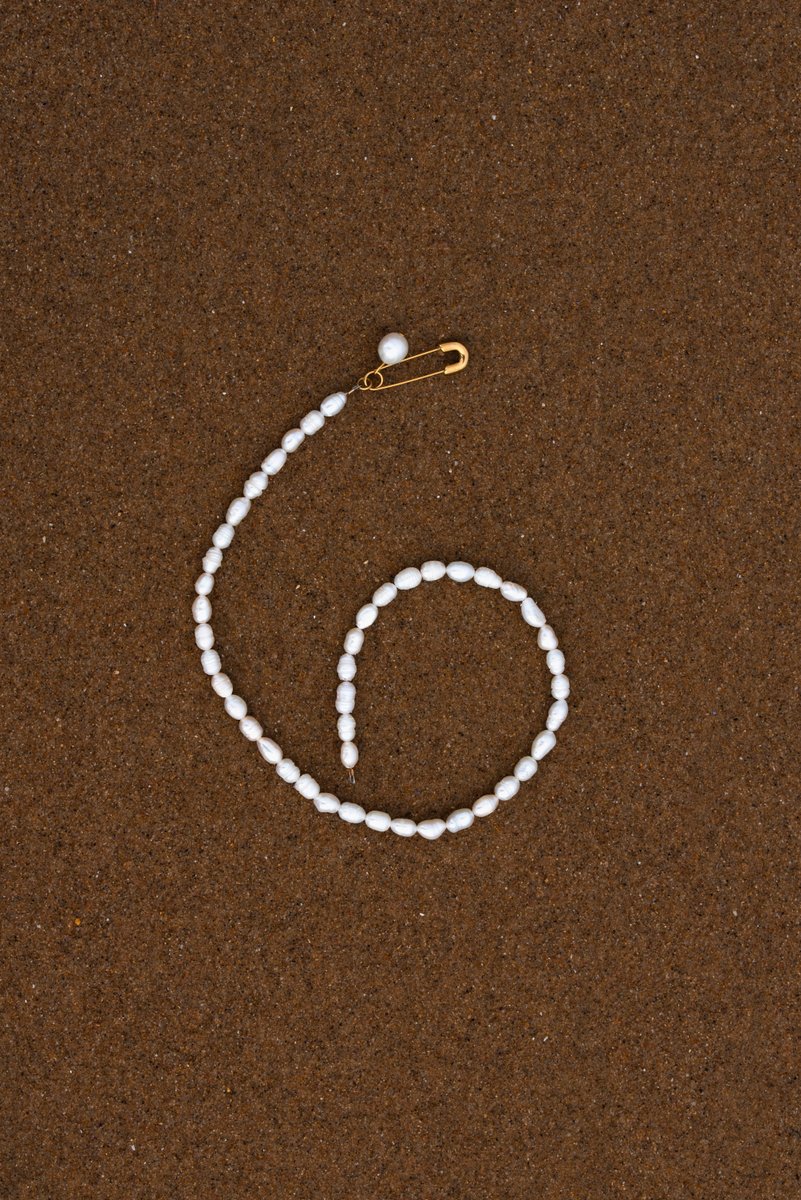 Pearl necklace arranged in a spiral shape on a smooth brown sandy surface, with a single safety pin clasp. The composition evokes a sense of fluidity and simplicity. by Sydney SG