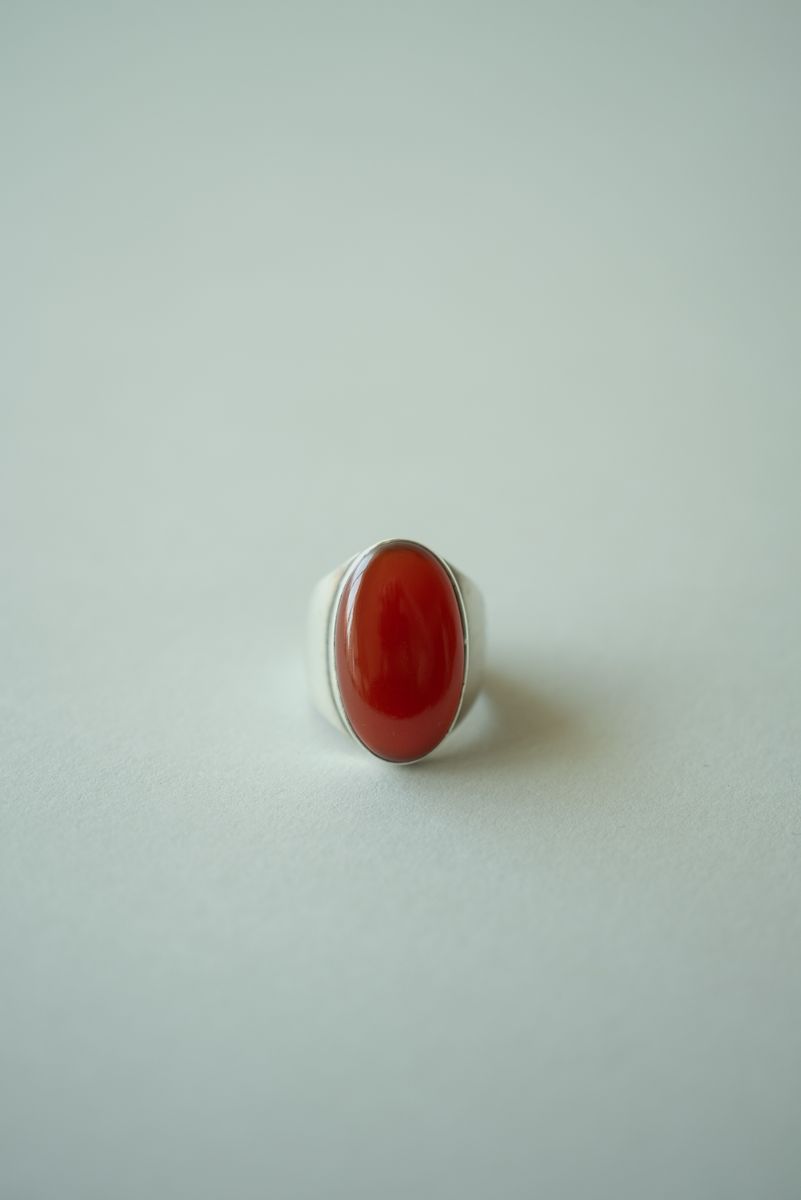 An elegant silver cocktail ring featuring a large oval red carnelian stone set in a minimalist bezel. The polished scarlet centerpiece reflects subtle light from its domed surface, creating a bold yet refined statement against the pale background. by Sydney SG
