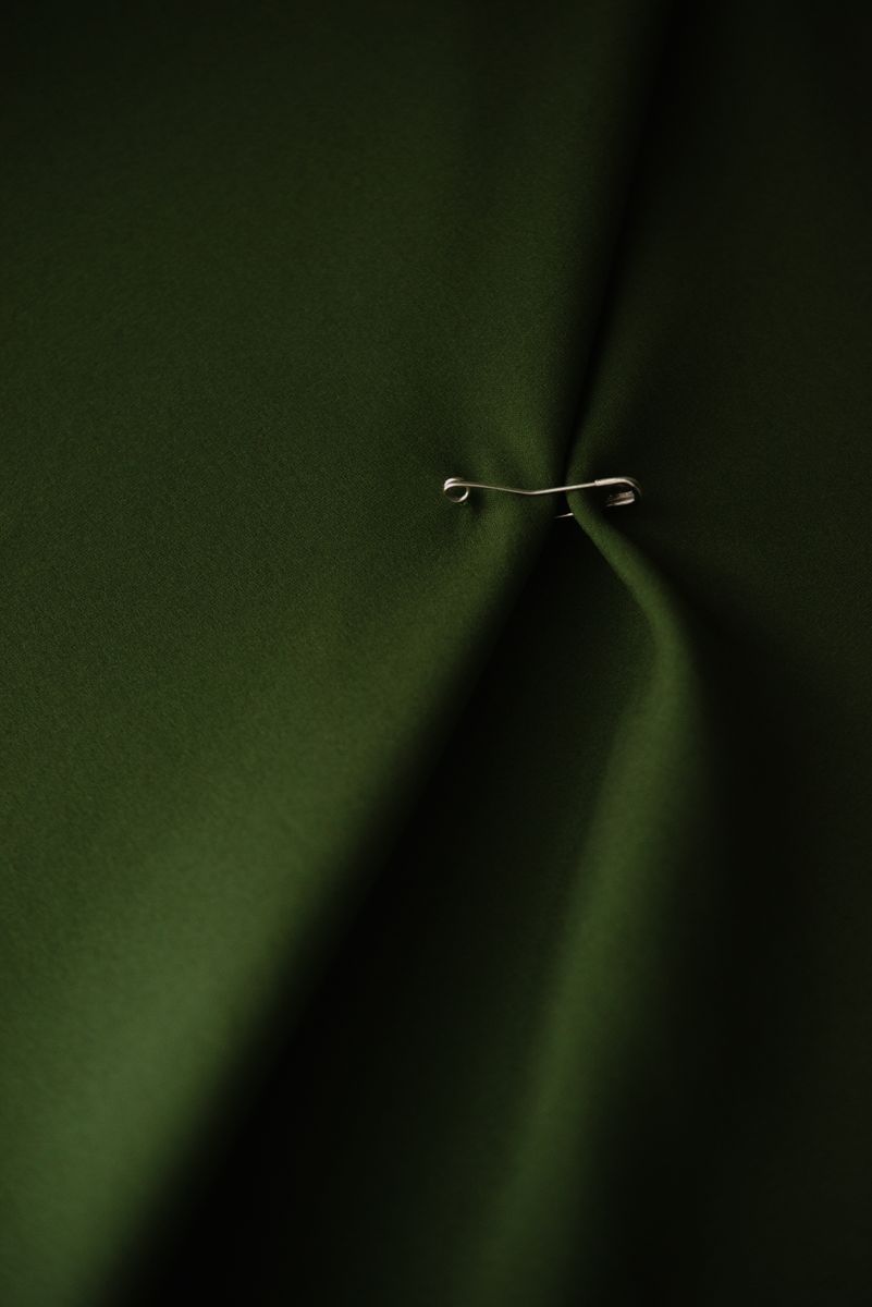 Macro view of a bent safety pin gently gathering folds of deep forest green fabric. The subtle interplay of light and shadow creates valleys in the textile, while the metallic pin offers a delicate focal point that speaks to both function and adornment. by Sydney SG