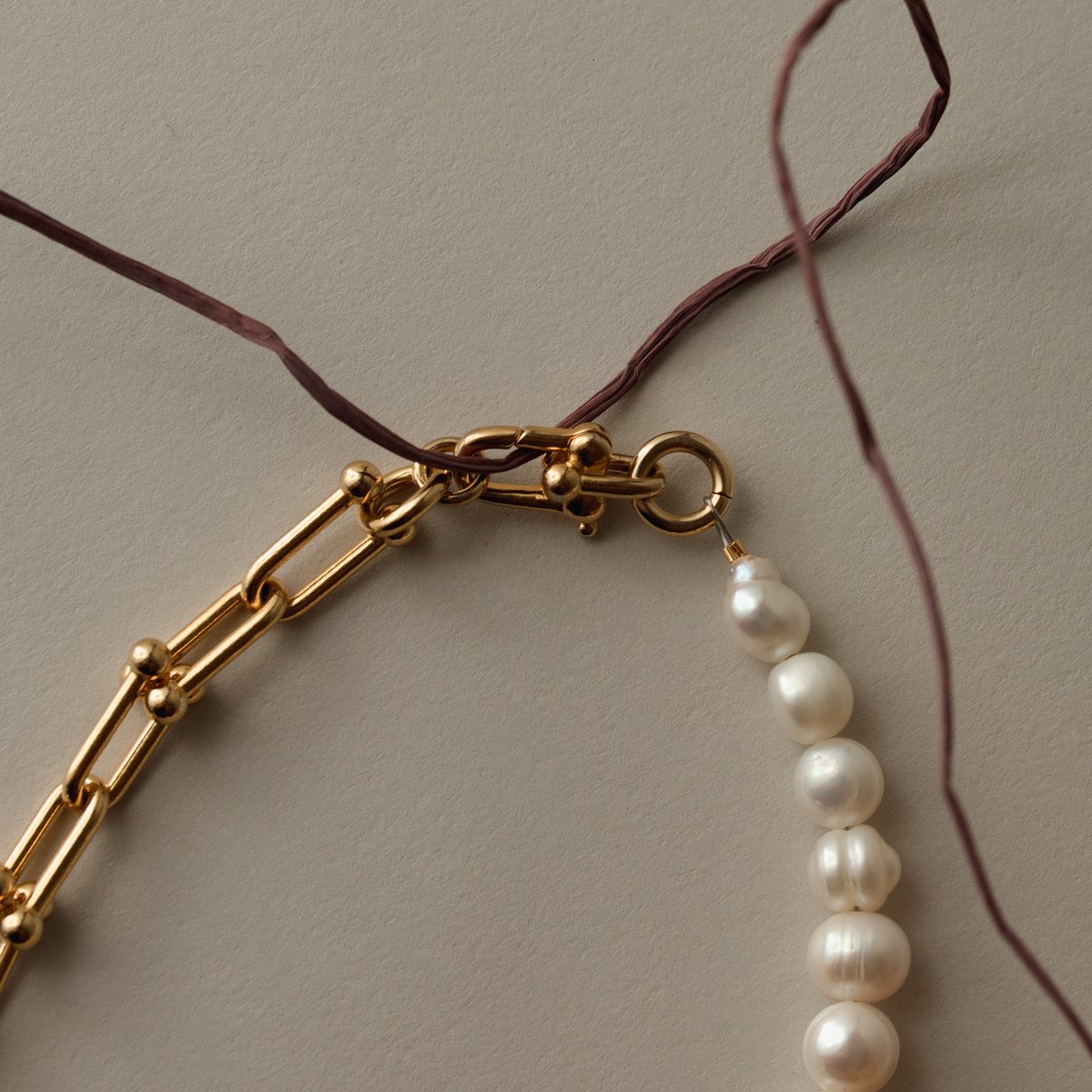 Close-up of a gold chain clasp attached to a string of pearls, looped around a thin brown plant stem. Minimalist and delicate jewelry arrangement on a neutral background. by Sydney SG