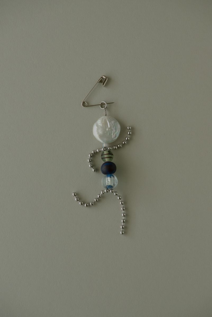 Delicate silver stick figure charm with a luminous pearl disc head and strategically placed beads in blue, striped, and clear glass forming the body. The curved chain limbs create a sense of movement while the safety pin connector suggests versatile wearability. by Sydney SG