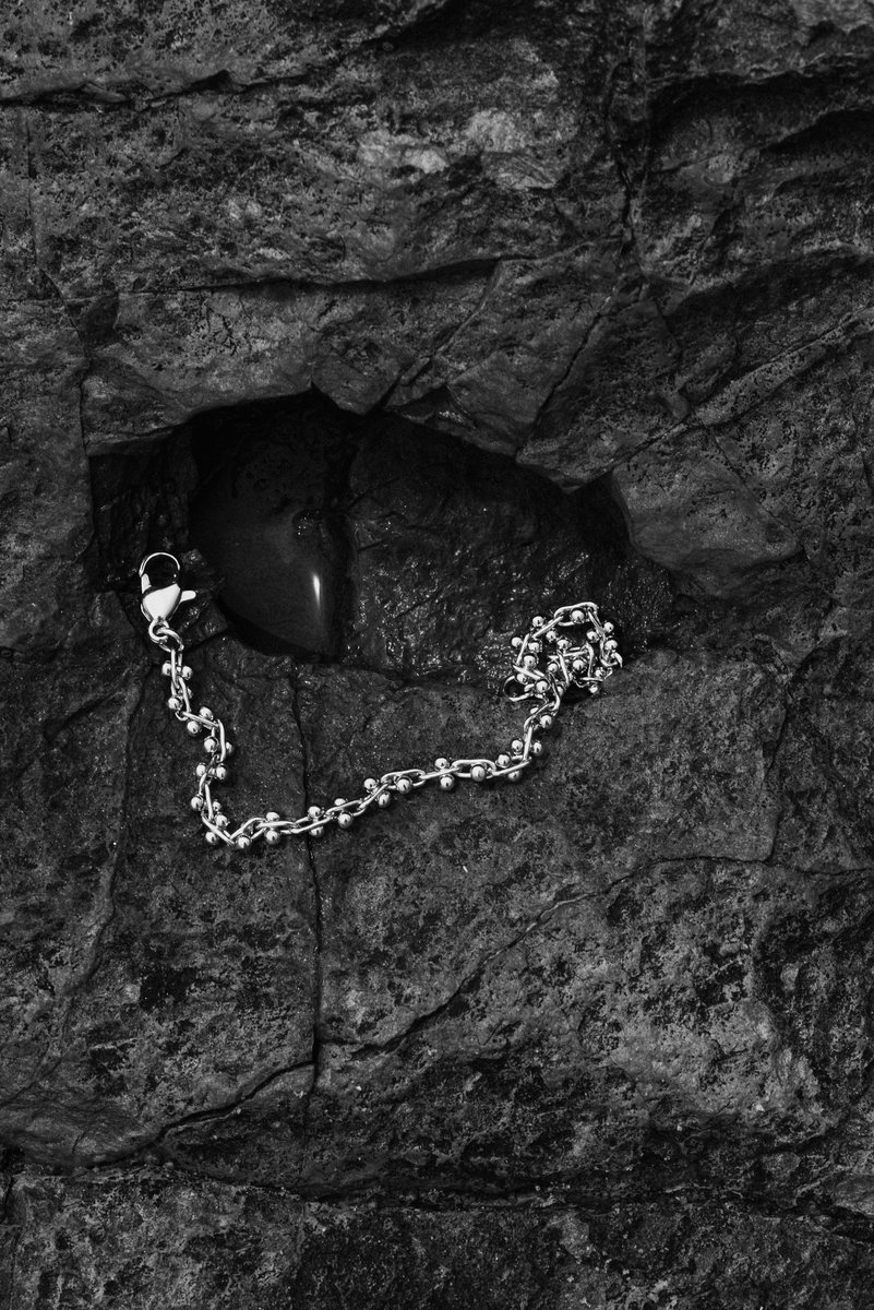 Silver chain laid across dark, rugged rocks with a small puddle of water. The contrast between the smooth metal and rough stone adds a dramatic, organic touch. by Sydney SG