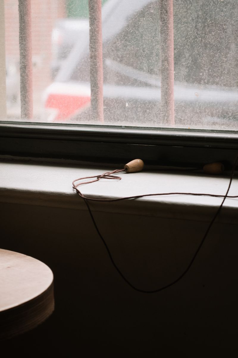 A wooden curtain pull with red-brown thread sits on a white windowsill with dust-covered glass visible behind it. The simple crafting object is framed by the textured, water-streaked window surface. by Sydney SG