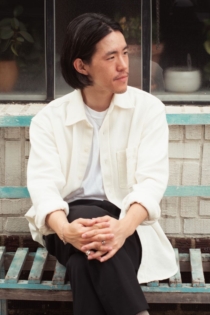A person in a white shirt sits calmly on weathered wooden steps with turquoise paint, hands folded. The composed portrait shows the subject in a moment of quiet reflection. by Sydney SG