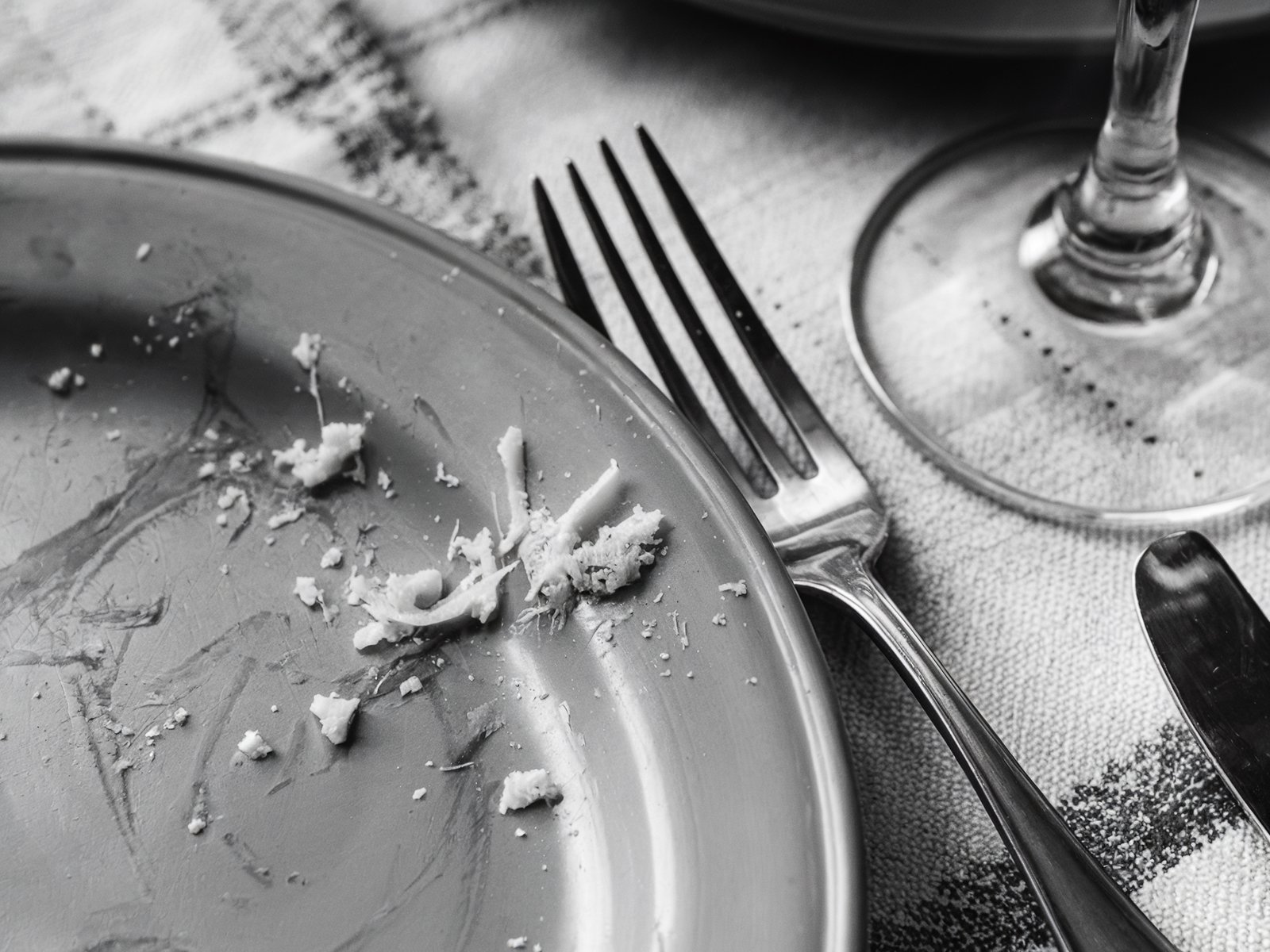 Black and white photo of an empty plate with cheese crumbs, fork, and a wine glass. A post-meal scene capturing the remnants of a casual meal. Image generated with AI. by Sydney SG