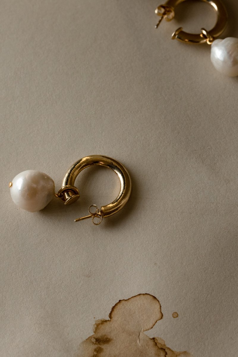 Single gold hoop earring with a white pearl placed on a neutral surface, next to a coffee stain. A minimalist still life with a rustic touch. by Sydney SG