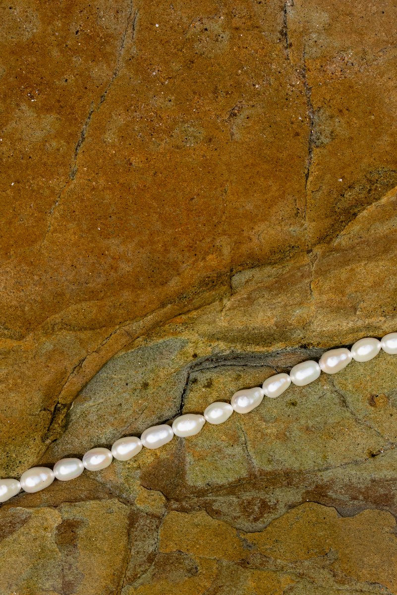 Pearl necklace placed diagonally against a rugged, earthy-toned rock surface. The smooth pearls contrast with the rough texture of the stone, creating an elegant, natural composition. by Sydney SG