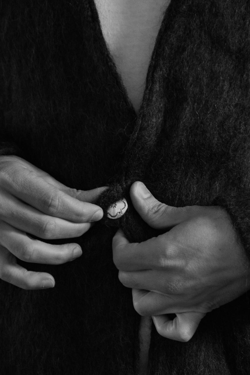 Intimate black and white closeup of hands fastening a decorative button on a dark wool garment. The gentle precision of the gesture and textural contrast between skin and fabric create a moment of quiet ritual. by Sydney SG
